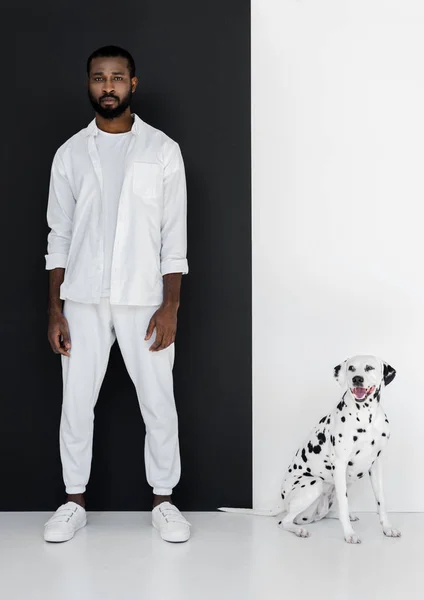 Bel homme afro-américain élégant en vêtements blancs et chien dalmate debout près du mur noir et blanc — Photo de stock