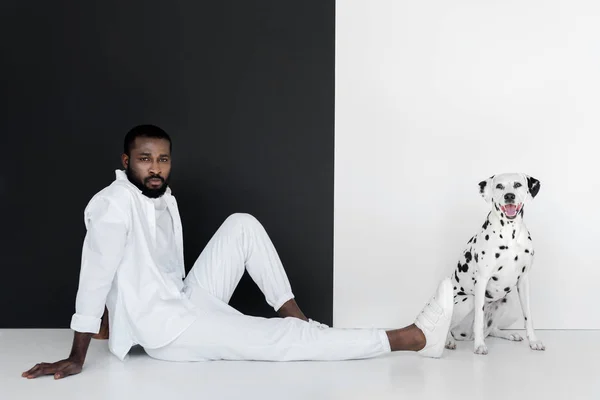 Bel homme afro-américain élégant en vêtements blancs et chien dalmate assis près du mur noir et blanc — Photo de stock