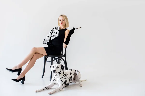 Jolie femme blonde élégante en robe noire assise sur une chaise, chien dalmate couché sur le sol — Photo de stock