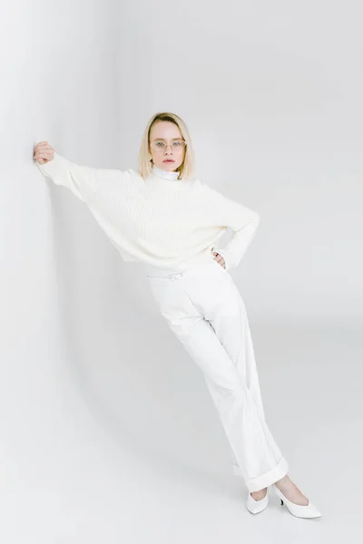 Beautiful stylish blonde woman in white clothes leaning on white wall and looking at camera — Stock Photo