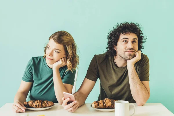Sonriente pareja usando smartphone y mirando hacia otro lado mientras desayunan juntos - foto de stock