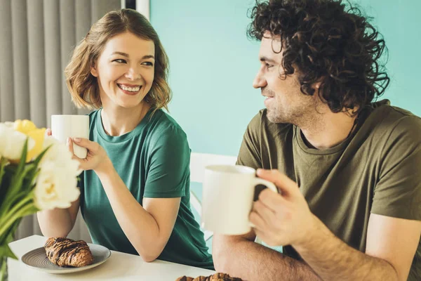 Feliz pareja sosteniendo tazas y sonriendo entre sí mientras desayunan juntos - foto de stock