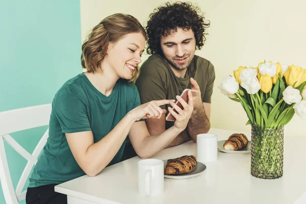 Smiling couple using smartphone while having breakfast together at home — Stock Photo