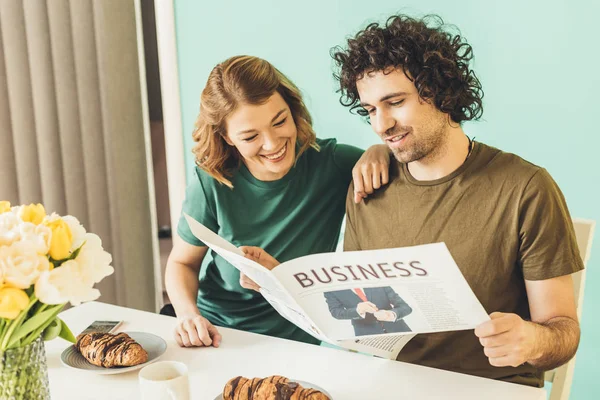 Happy couple in t-shirts reading newspaper while having breakfast together at home — Stock Photo