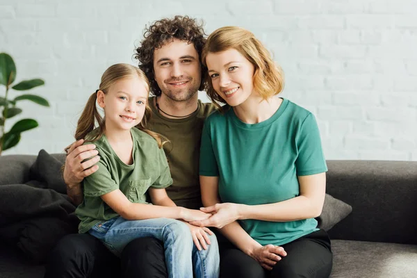 Padres felices con linda hijita sentada en el sofá y sonriendo a la cámara - foto de stock