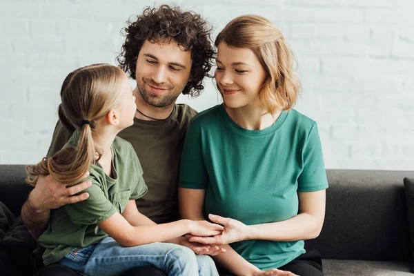 Padres felices mirando a la pequeña hija linda mientras está sentado en el sofá en casa - foto de stock