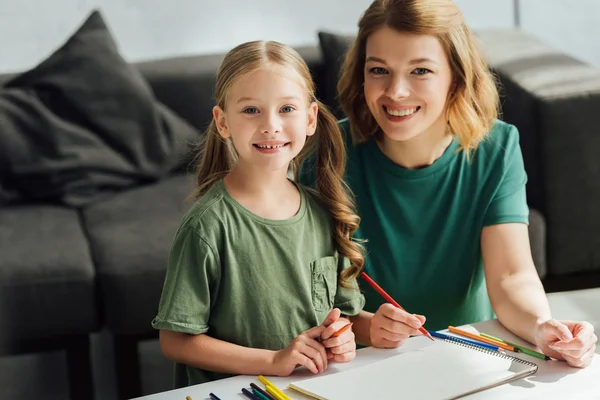 Счастливая мать и дочь рисуют цветными карандашами и улыбаются в камеру — стоковое фото