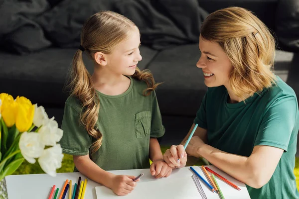 Mãe feliz e filha desenhando juntos e sorrindo uns aos outros em casa — Fotografia de Stock