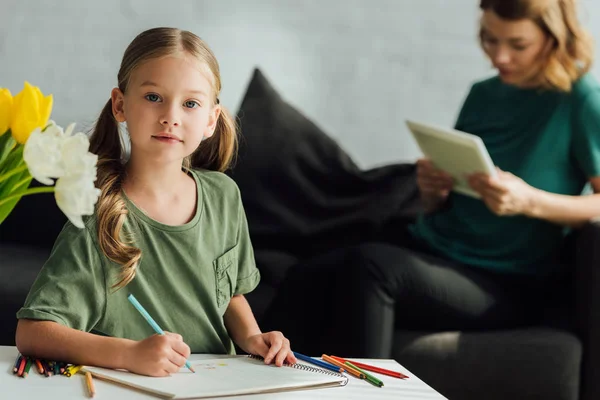 Niedliches kleines Kind zeichnet mit Buntstiften und schaut in die Kamera, während die Mutter das digitale Tablet hinter sich hat — Stockfoto