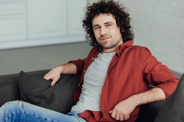 Retrato de un hombre guapo con el pelo rizado sentado en el sofá y mirando a la cámara en casa - foto de stock