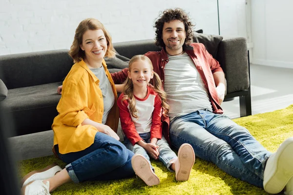 Happy young parents with cute little daughter sitting together on carpet and smiling at camera — Stock Photo