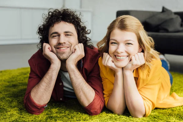 Hermosa feliz pareja acostada en la alfombra y sonriendo a la cámara en casa - foto de stock