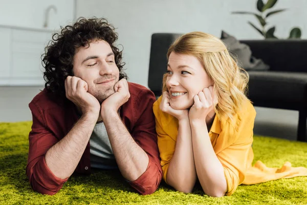 Hermosa feliz pareja acostada en la alfombra y sonriendo el uno al otro en casa - foto de stock