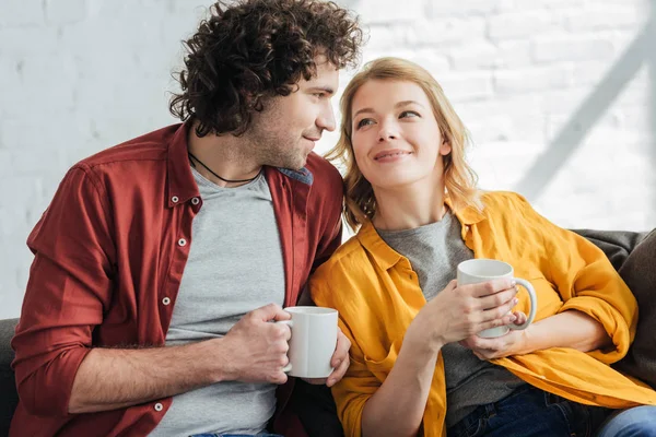 Glückliches junges Paar hält Tassen in der Hand und lächelt sich zu Hause an — Stockfoto
