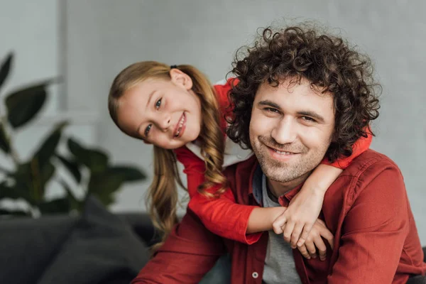 Linda hija pequeña abrazando feliz padre y sonriendo a la cámara en casa - foto de stock