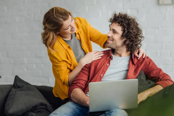 Glückliches junges Paar lächelt einander an, während es zu Hause Laptop benutzt — Stockfoto