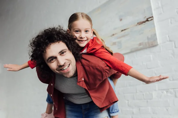 Feliz padre piggybacking linda hija pequeña y sonriendo a la cámara en casa - foto de stock