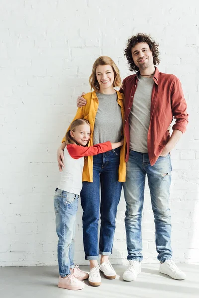 Vue pleine longueur des parents heureux avec fille embrassant et souriant à la caméra — Photo de stock