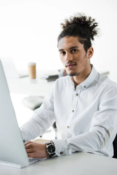 Joven hombre de negocios afroamericano trabajando con computadora de escritorio y mirando a la cámara - foto de stock