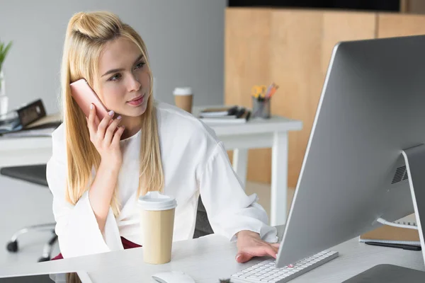 Schöne junge Geschäftsfrau, die mit dem Smartphone spricht und im Büro Desktop-Computer benutzt — Stockfoto