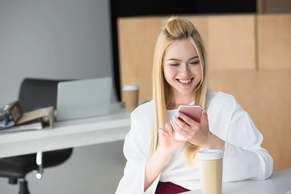 Hermosa mujer joven sonriente usando teléfono inteligente en el lugar de trabajo - foto de stock