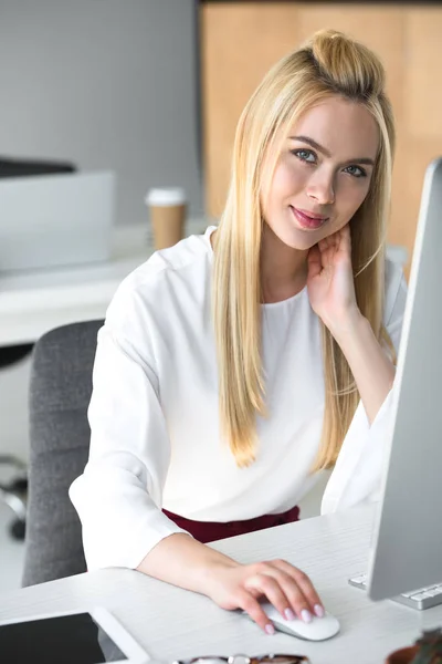 Bela jovem mulher usando computador desktop e sorrindo para a câmera no escritório — Fotografia de Stock
