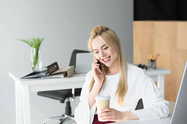 Smiling young businesswoman holding disposable coffee cup and talking by smartphone in office — Stock Photo