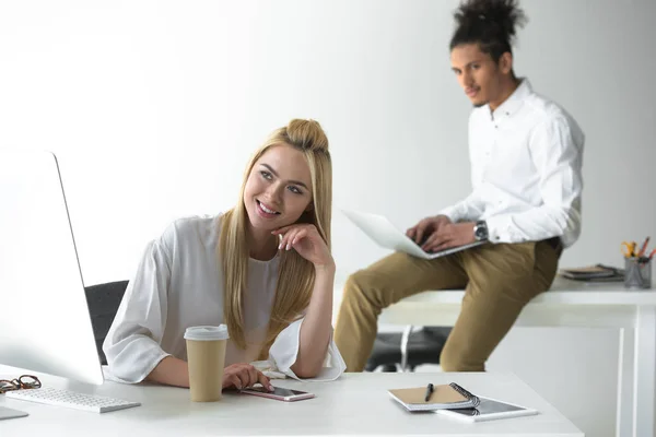 Atraente sorrindo jovem mulher sentada no local de trabalho e empresário afro-americano usando laptop atrás — Fotografia de Stock