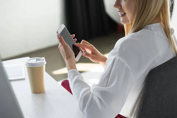 Recortado disparo de mujer joven sonriente usando teléfono inteligente en el lugar de trabajo - foto de stock