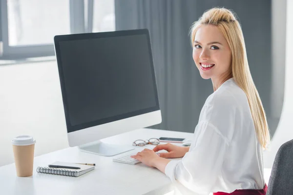 Красивая молодая деловая женщина улыбается в камеру во время работы с настольным компьютером в офисе — стоковое фото