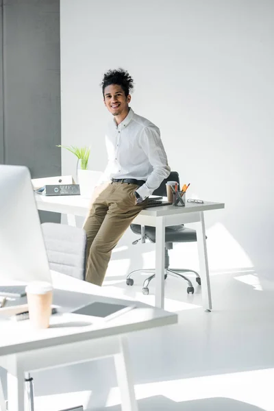 Jovem empresário afro-americano bonito sentado na mesa e sorrindo para a câmera no escritório — Fotografia de Stock