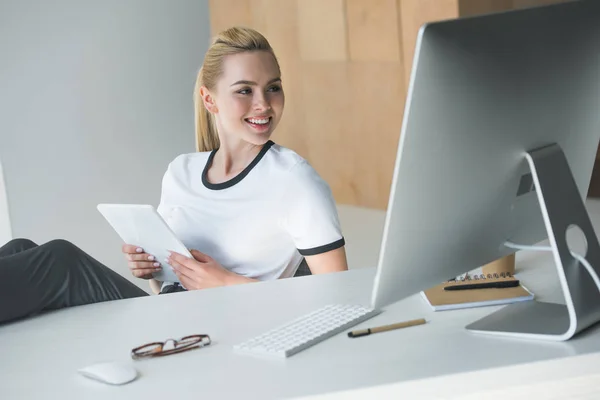 Красива молода жінка тримає цифровий планшет і дивиться на настільний комп'ютер в офісі — стокове фото