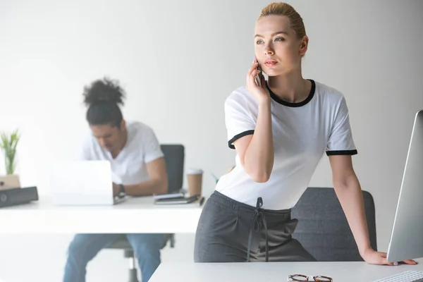 Schöne junge Frau, die mit dem Smartphone spricht und wegschaut, während sie sich im Büro an den Tisch lehnt — Stockfoto