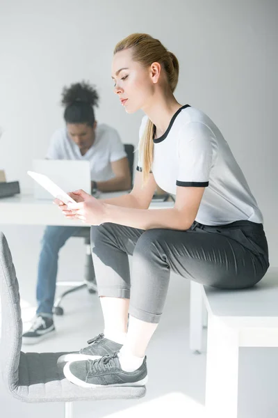 Atractiva joven sentada en la mesa y usando tableta digital en la oficina - foto de stock