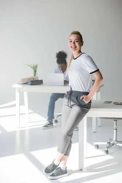 Fröhliche junge Frau lehnt am Tisch und lächelt in die Kamera im Büro — Stockfoto