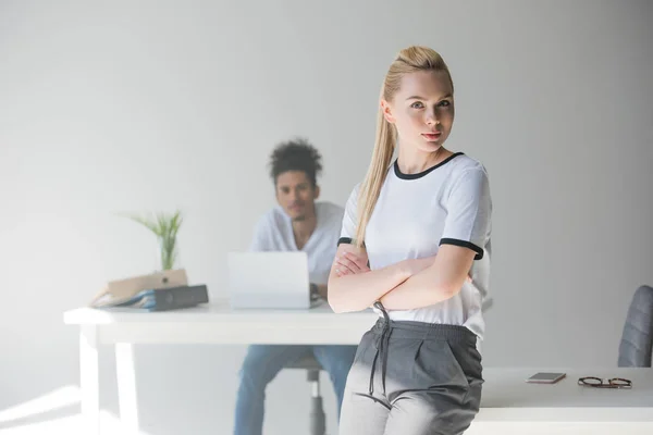 Jovem mulher confiante de pé com os braços cruzados e olhando para a câmera enquanto colega do sexo masculino trabalhando atrás no escritório — Fotografia de Stock