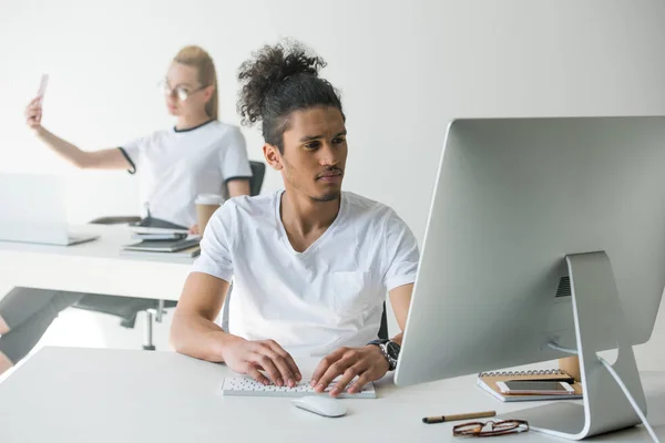 Junger afrikanisch-amerikanischer Mann benutzt Desktop-Computer und Kollegin macht Selfie im Büro — Stockfoto