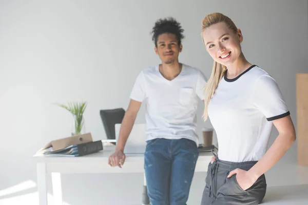 Belle jeune femme souriant à la caméra tout en se tenant avec un collègue afro-américain au bureau — Photo de stock