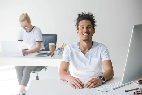 Junger afrikanisch-amerikanischer Mann lächelt in die Kamera, während er im Büro mit einer Kollegin im Hintergrund arbeitet — Stockfoto