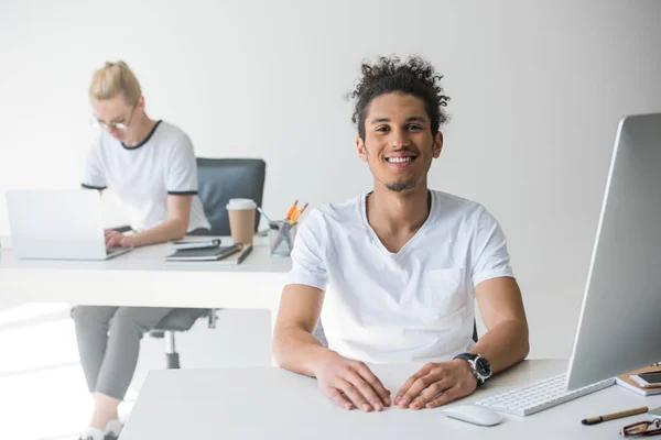 Joven afroamericano hombre sonriendo en cámara mientras trabajando en oficina con mujer colega detrás - foto de stock