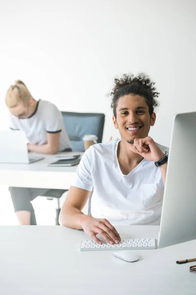 Bonito jovem afro-americano homem usando computador desktop e sorrindo para a câmera no escritório — Fotografia de Stock