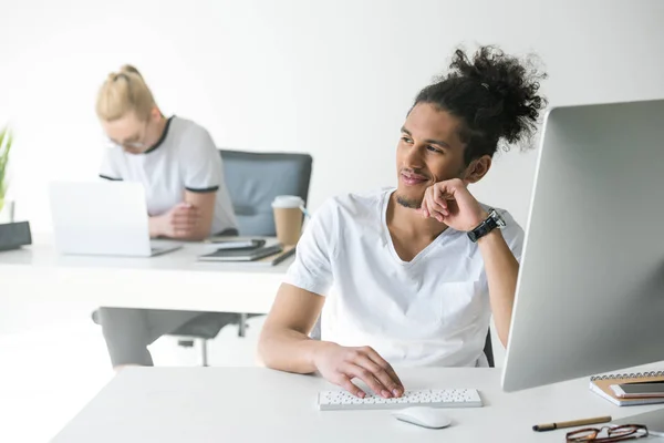 Lächelnder junger afrikanisch-amerikanischer Mann benutzt Desktop-Computer und schaut im Büro weg — Stockfoto