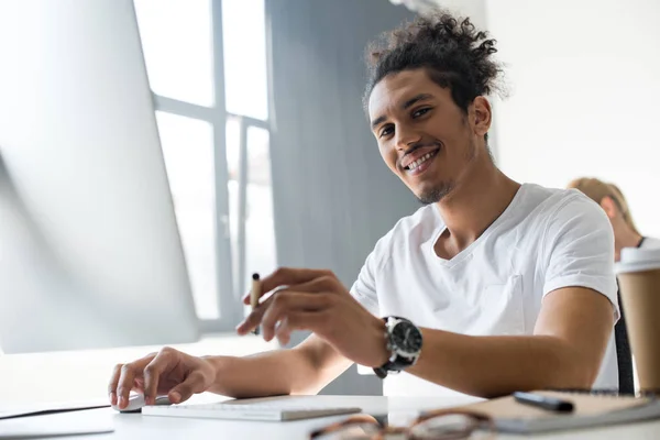 Schöner junger afrikanisch-amerikanischer Mann lächelt in die Kamera, während er im Büro mit dem Desktop-Computer arbeitet — Stockfoto