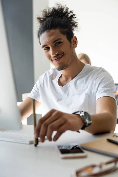 Joven afroamericano hombre sosteniendo pluma y sonriendo a la cámara mientras está sentado en el lugar de trabajo - foto de stock