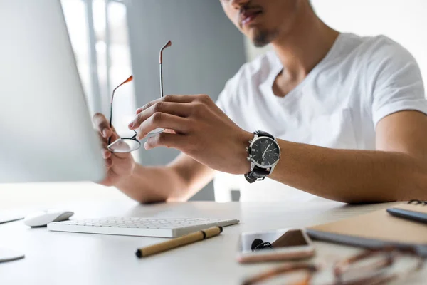 Tiro cortado de jovem segurando óculos no local de trabalho — Fotografia de Stock