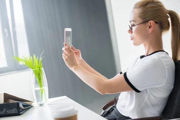 Вид сбоку красивой молодой блондинки, делающей селфи со смартфоном в офисе — стоковое фото