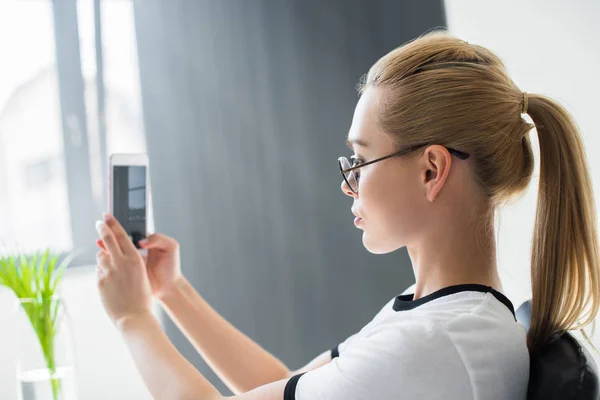 Vista lateral de la joven rubia tomando selfie con teléfono inteligente en la oficina — Stock Photo