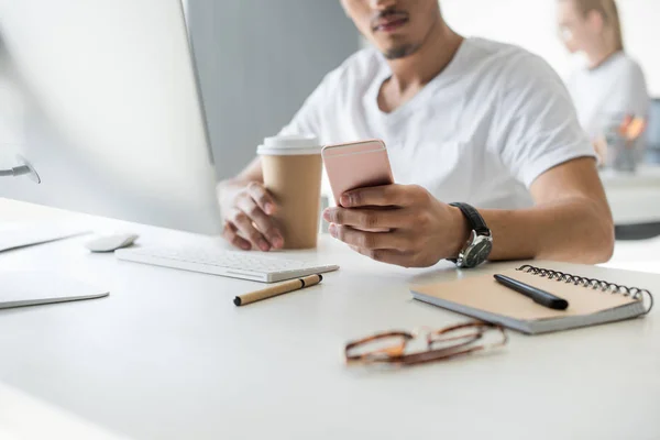 Cropped shot of young african american man using smartphone and holding disposable coffee cup at workplace — Stock Photo