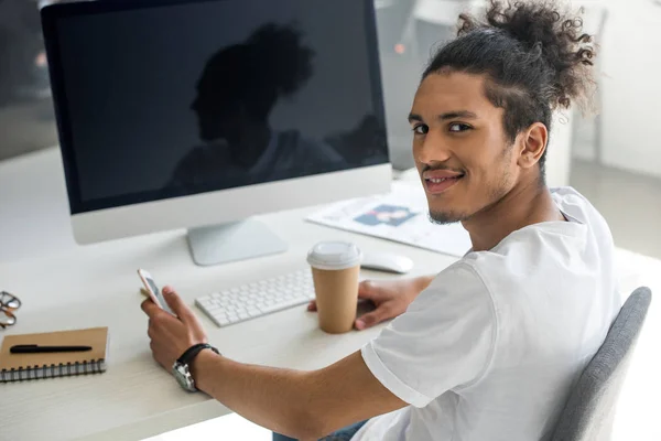 Young african american man with paper cup and smartphone smiling at camera at workplace — Stock Photo