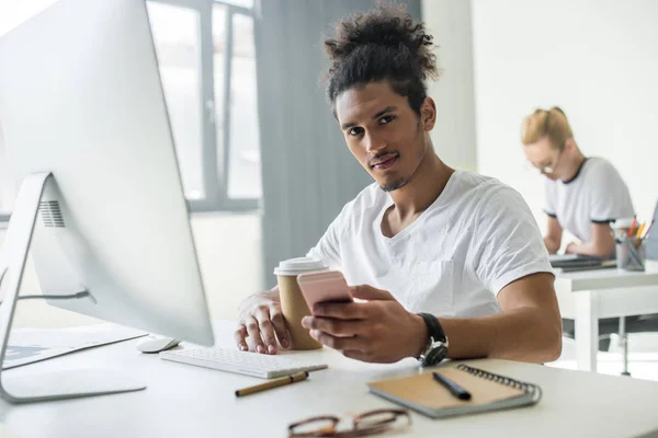 Jeune homme afro-américain regardant la caméra tout en utilisant un smartphone et un ordinateur de bureau au bureau — Photo de stock